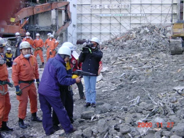 Group of rescue workers and officials in helmets and protective gear inspecting debris and rubble at a construction site or disaster zone with damaged structures in the background.