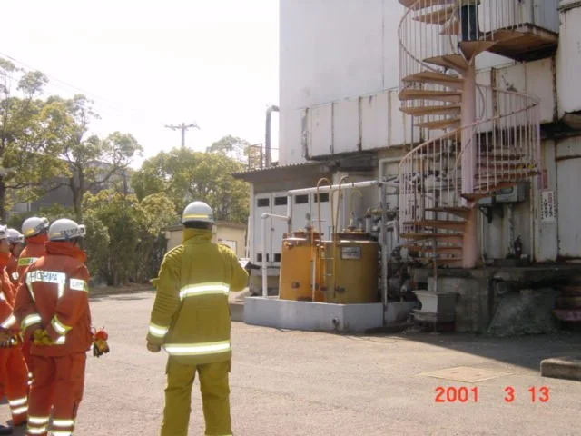 Group of firefighters in orange and yellow uniforms standing outside a building with a fire escape staircase, near equipment, on a sunny day.