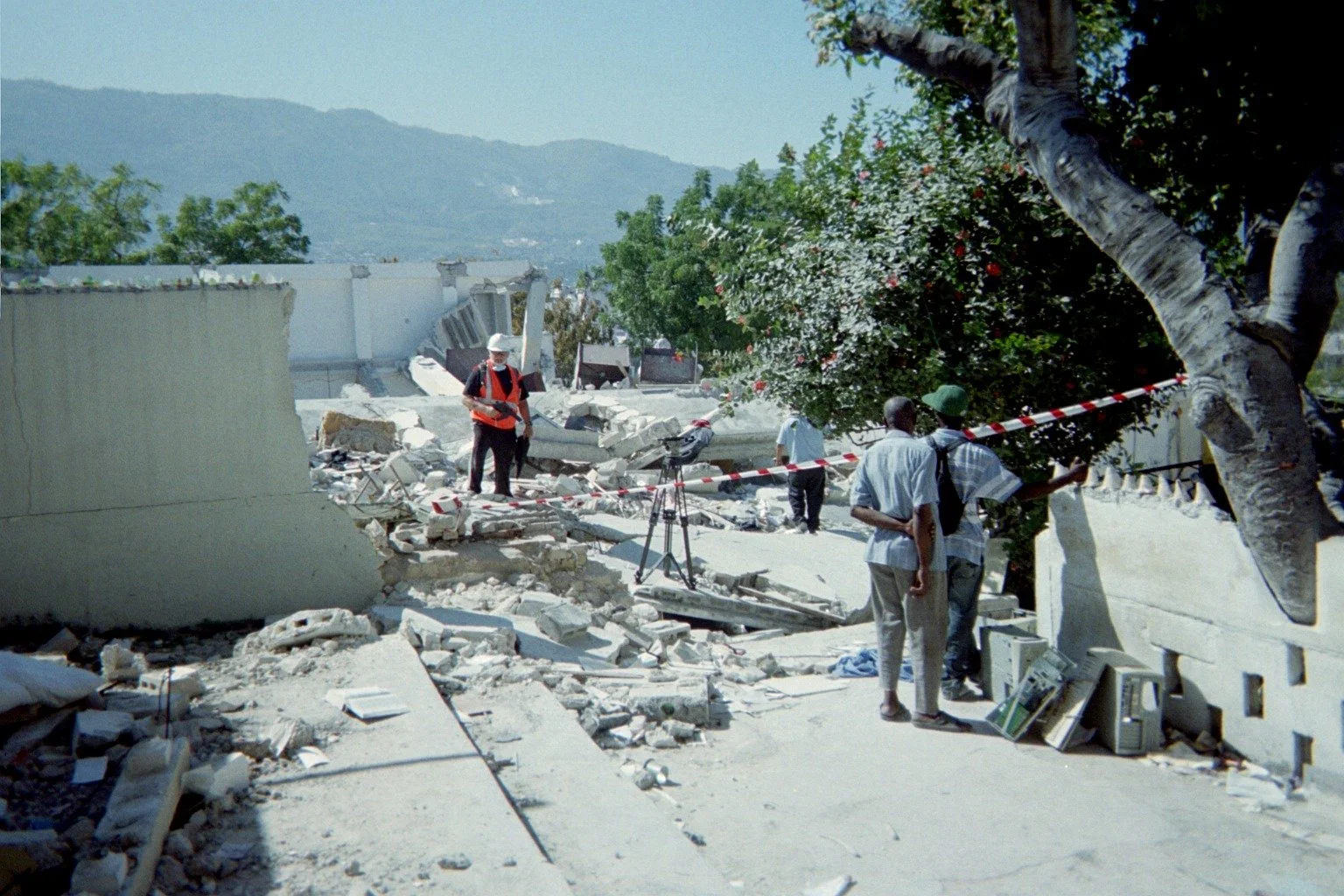 Scene of building destruction with debris, a fallen tree, and several men inspecting the damage, some taking photos, in an outdoor area with mountains in the background.