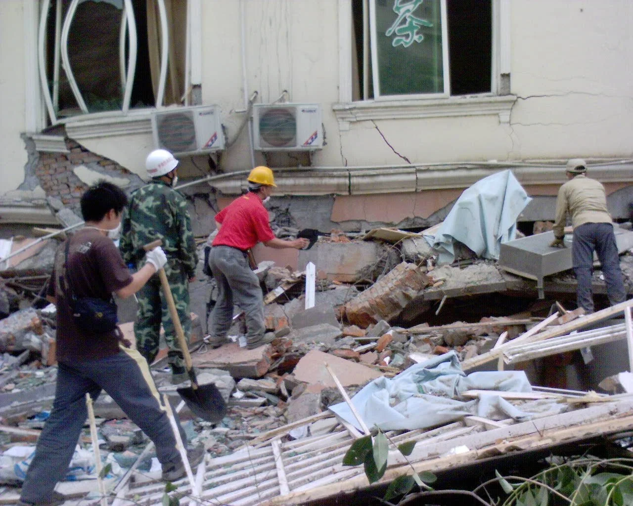Workers in helmets and masks clearing debris from a collapsed building with a damaged wall and exposed air conditioning units.