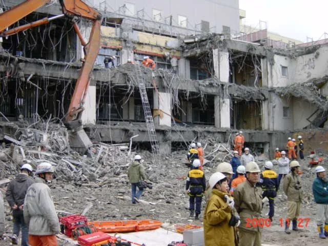 Construction workers and emergency responders in safety gear at a building collapse site with debris and damaged structure.