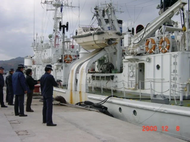 A group of men standing on a dock next to a large white naval ship with crew members on deck, equipped with various radar and communication antennas, life rings, and a large crane, with a cloudy sky in the background.