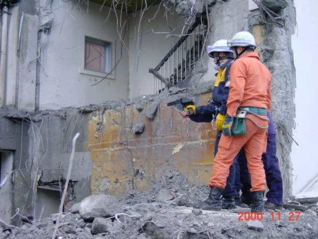 Three construction workers in safety gear and helmets inspecting a damaged building with debris and exposed concrete.