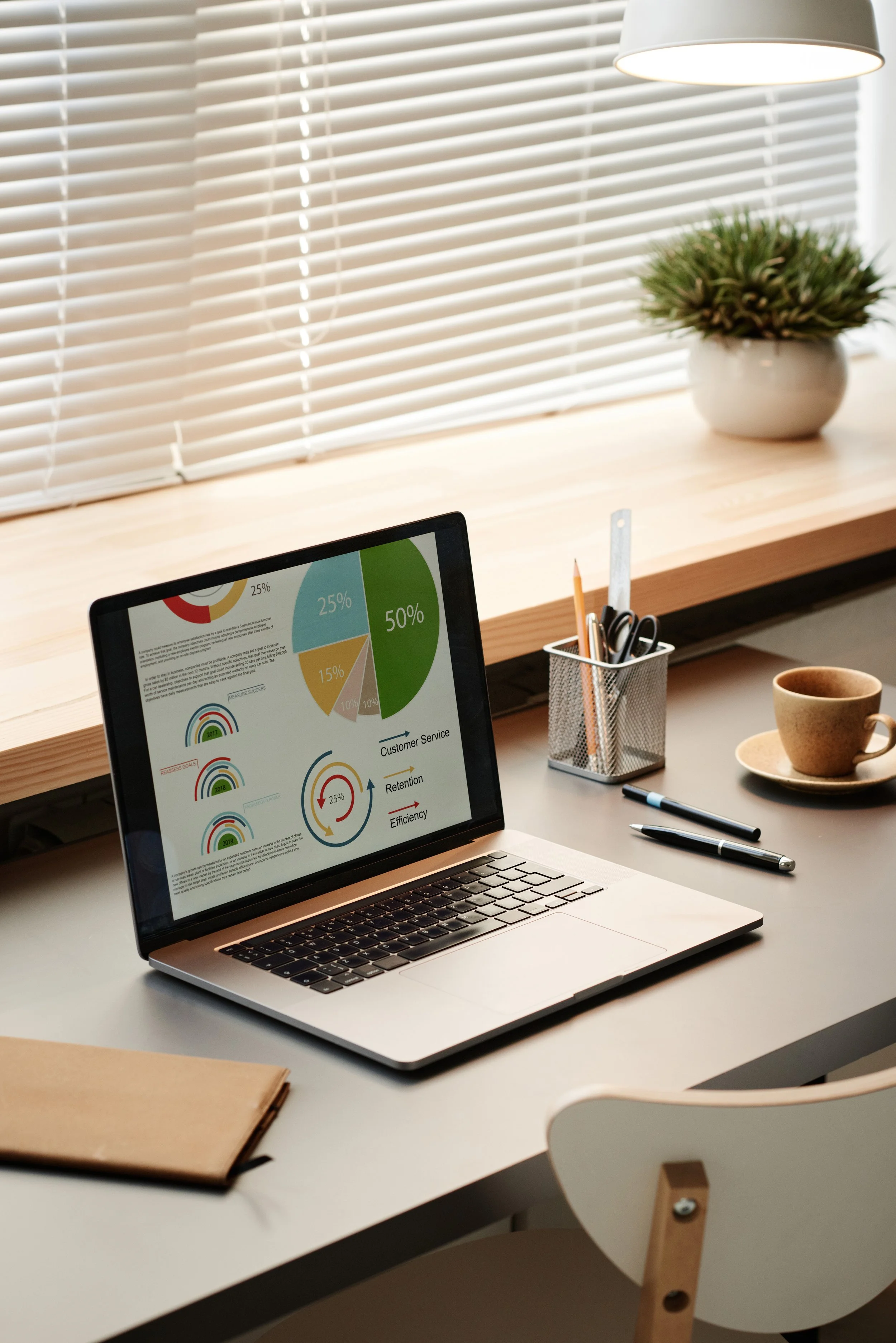 Work desk with a laptop displaying colorful charts and graphs, a notebook, pens, a cup of coffee, a pen holder, a window with blinds, and a potted plant.