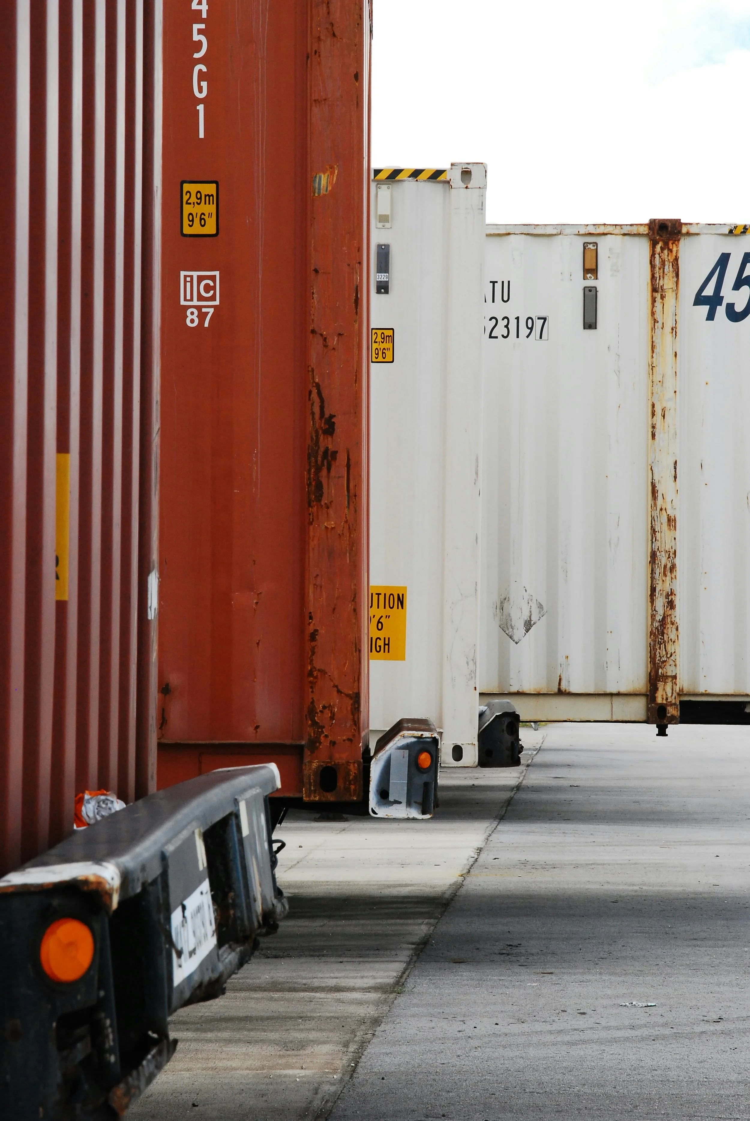 Several shipping containers in red, white, and beige colors lined up on a concrete ground, some showing signs of rust and wear, with low-angle view.