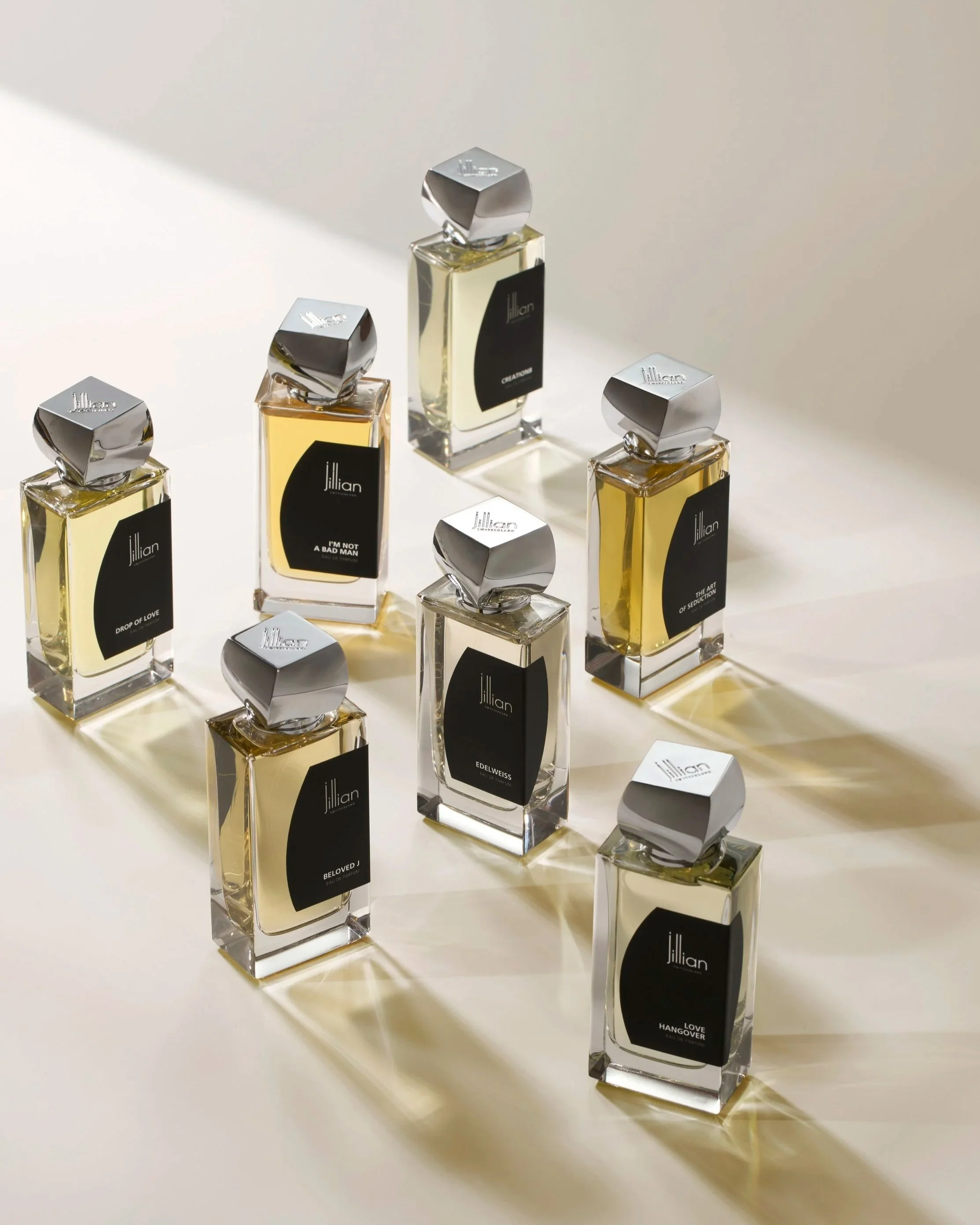 Seven bottles of Jillian perfume with black labels and silver caps arranged on a white surface, casting shadows.
