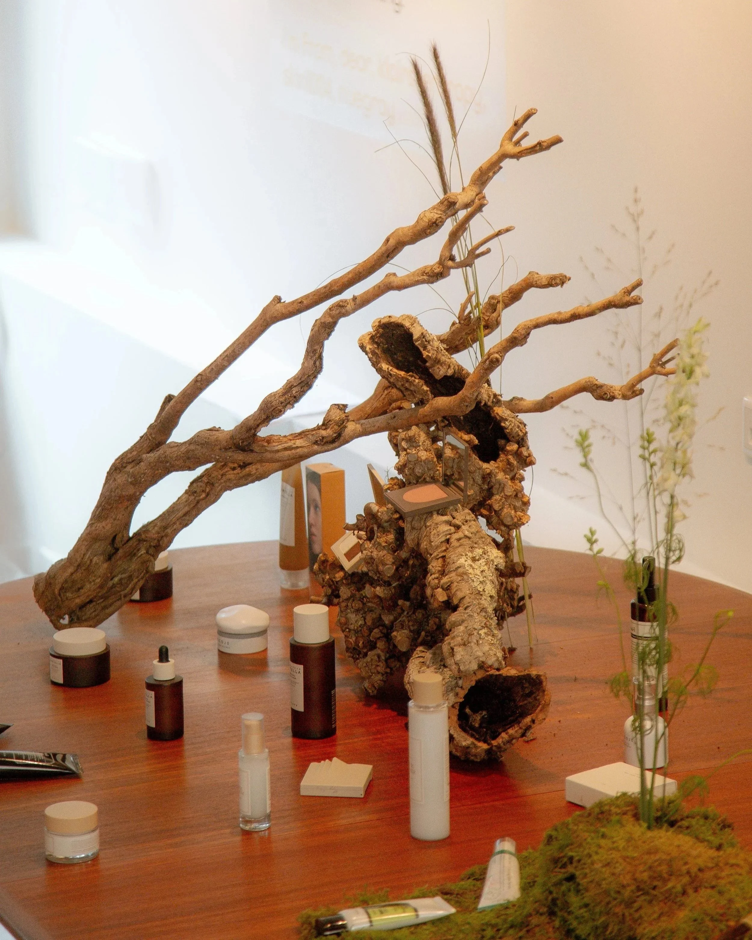 A wooden table displaying skincare products, a glass spray bottle, a small white item, and decorative natural elements including driftwood, bark, and greenery.