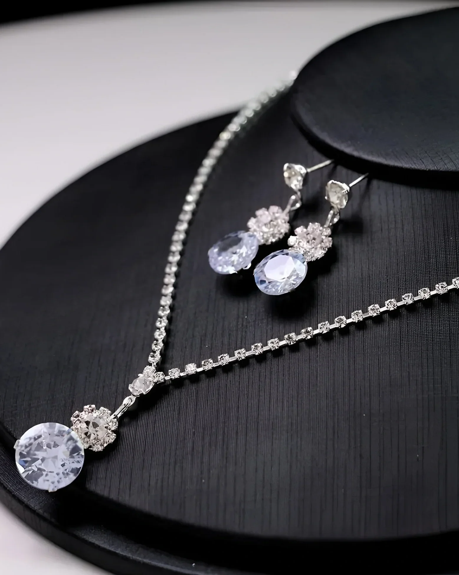 Set of jewelry including a necklace, earrings, and a bracelet, all adorned with clear gemstones, displayed on a black circular surface.