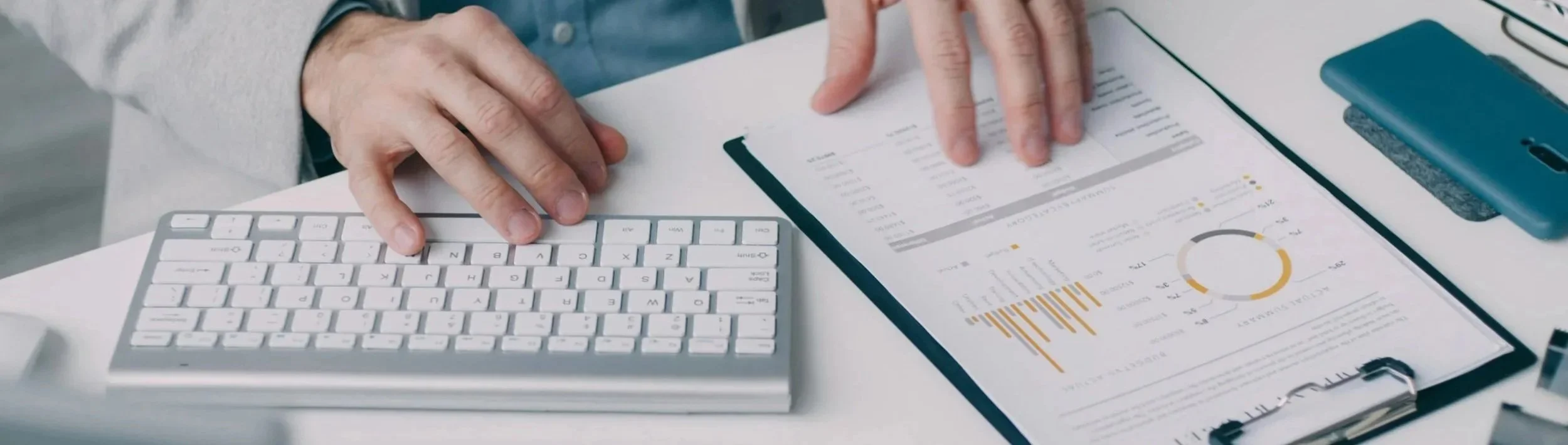 Person using a white keyboard and analyzing financial documents with charts and graphs on a clipboard.