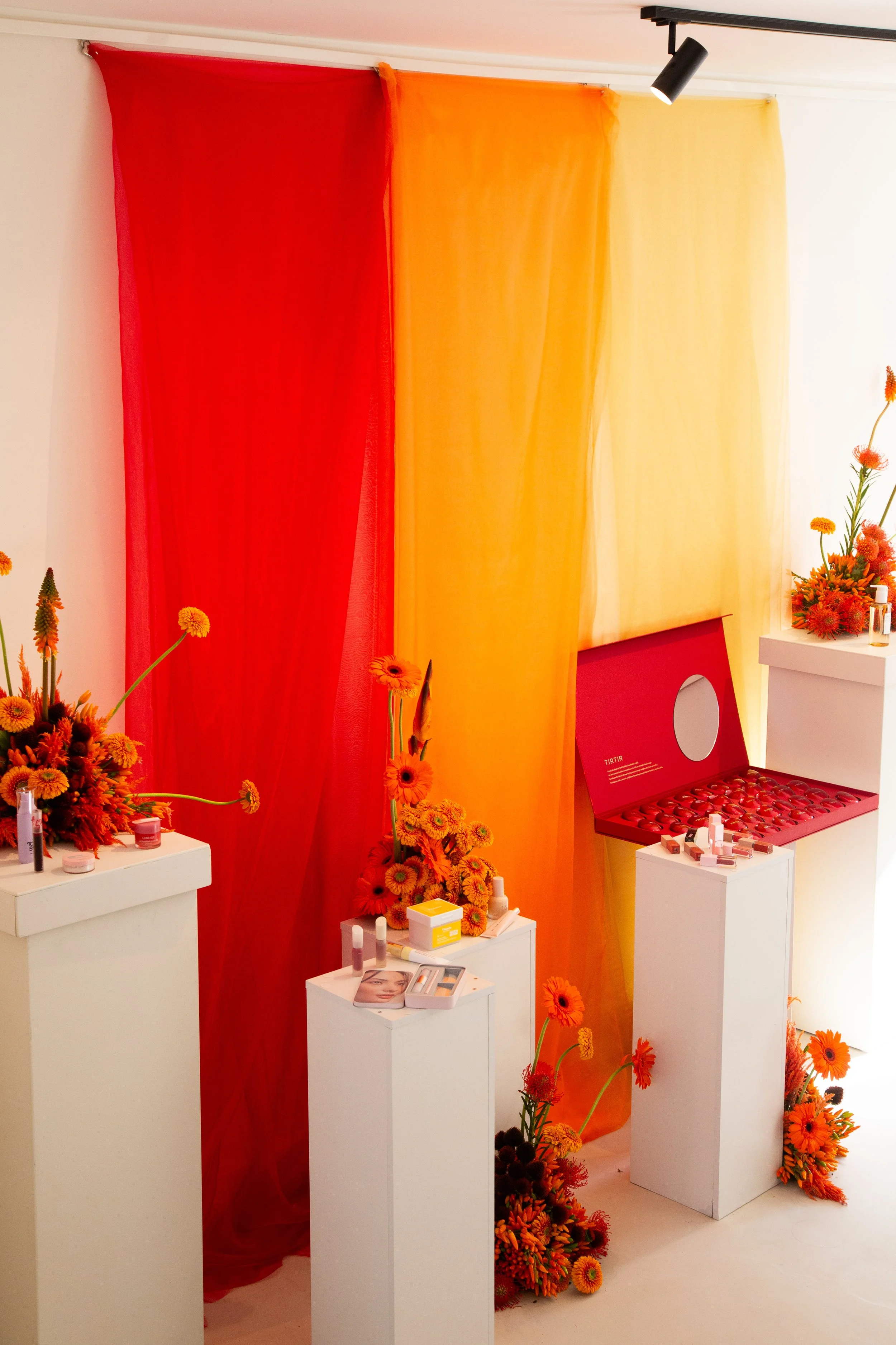 Display with red, orange, and yellow fabric backdrops, white pedestals with beauty products, orange flowers, and a red box filled with small bottles, in a brightly lit room.