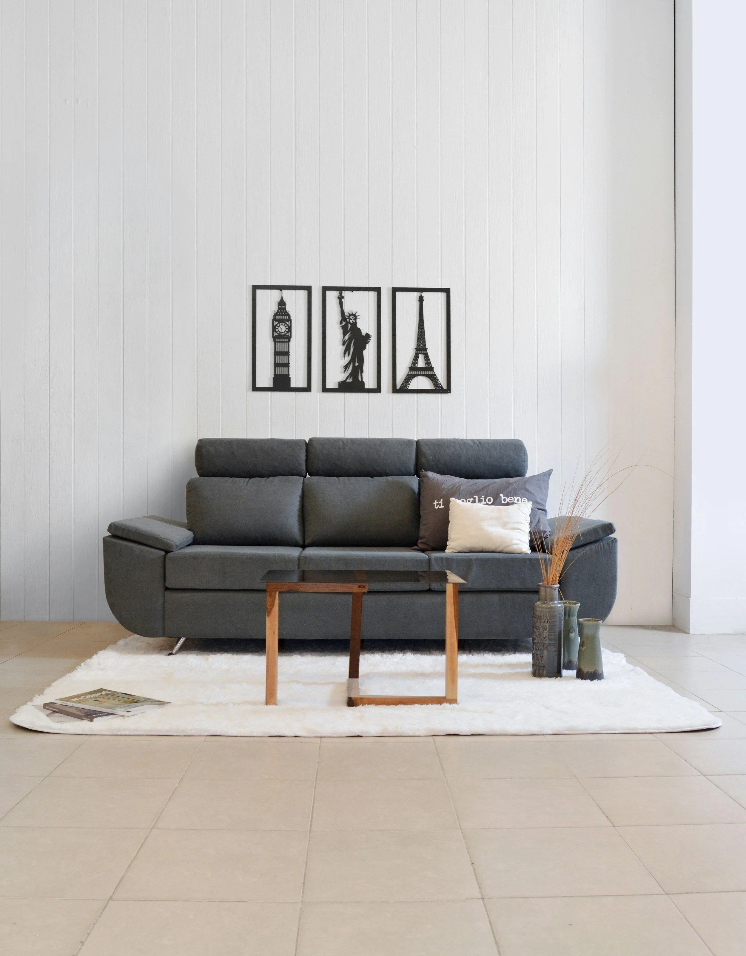 Living room featuring a gray sofa with pillows, a small wooden coffee table, a white shag rug, decorative vases with dried plants, and black framed wall art of Big Ben, Statue of Liberty, and Eiffel Tower.