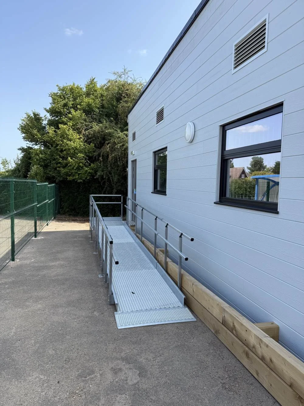 A portable metal ramp with handrails leading to a building entrance, with a wooden planter border along the building wall and trees in the background.