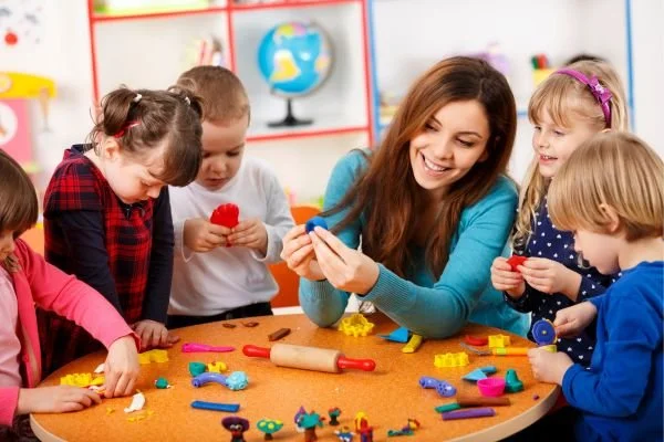 A female teacher and five children playing with colorful clay and modeling tools at a classroom table.