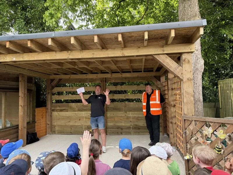A man speaking to a group of children outdoors, with another man in a high-visibility vest standing nearby. The scene is under a wooden structure with a leaf-covered tree in the background.