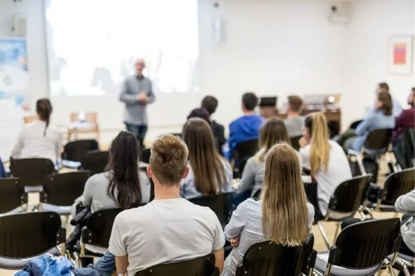 A classroom with students seated facing a teacher standing at the front.
