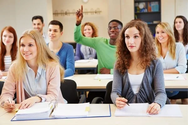 College classroom with students attentively listening, some taking notes, and one student raising hand