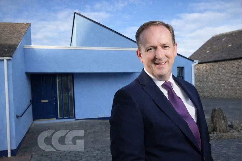 A man in a dark suit, white shirt, and purple tie standing outdoors in front of a modern blue building with an angled roof.