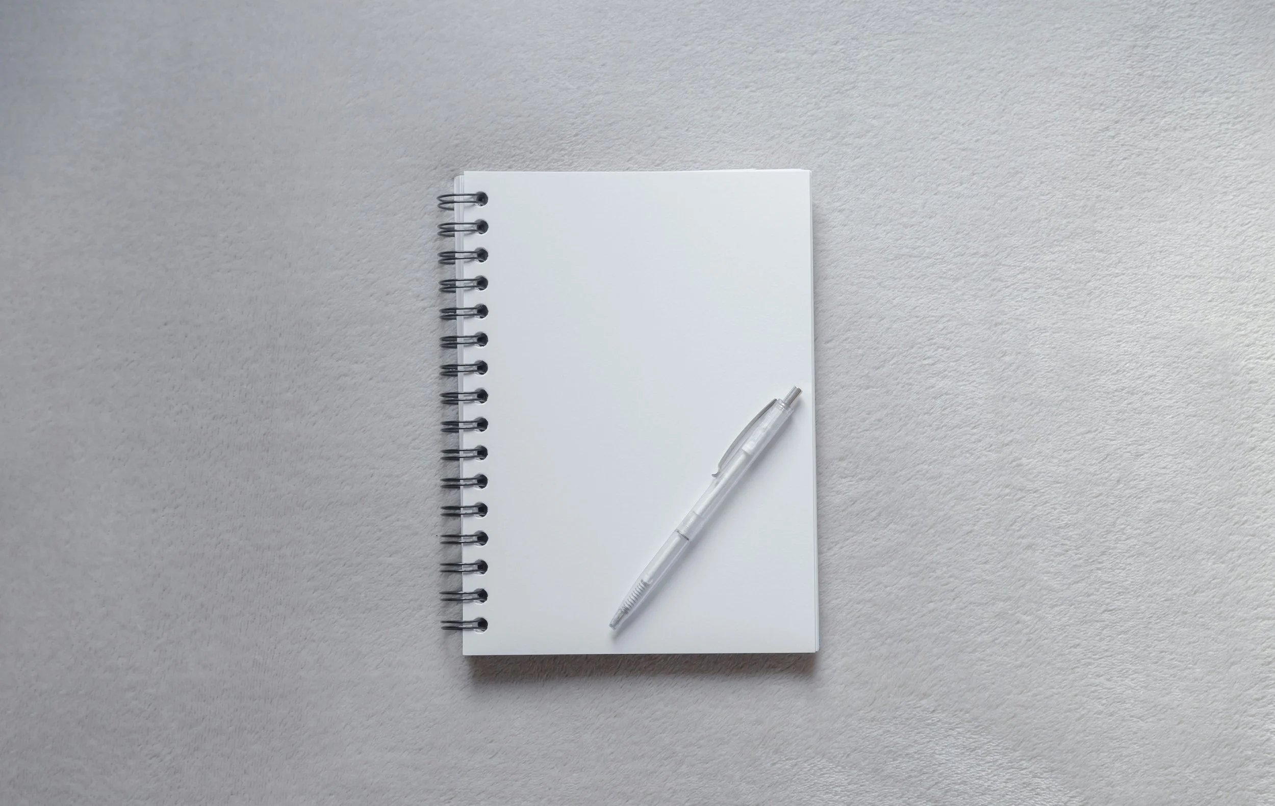 White spiral notebook with a transparent ballpoint pen resting on it, placed on a textured light gray surface.