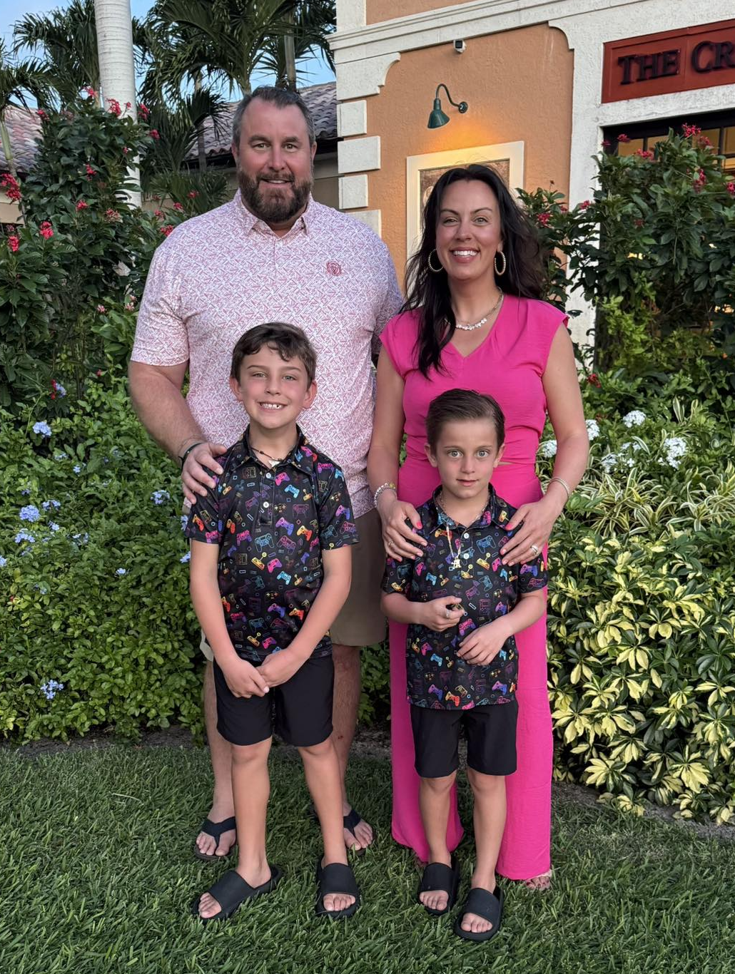 A family of four standing outdoors in front of a garden with bushes and flowers, in front of a tan-colored building. The father and mother are standing behind two young boys, all smiling at the camera. The father is tall with a beard, wearing a pink patterned shirt. The mother has long dark hair, wearing a bright pink dress with jewelry. The boys are wearing matching black shirts with colorful game controllers and shorts, with one boy slightly taller than the other.