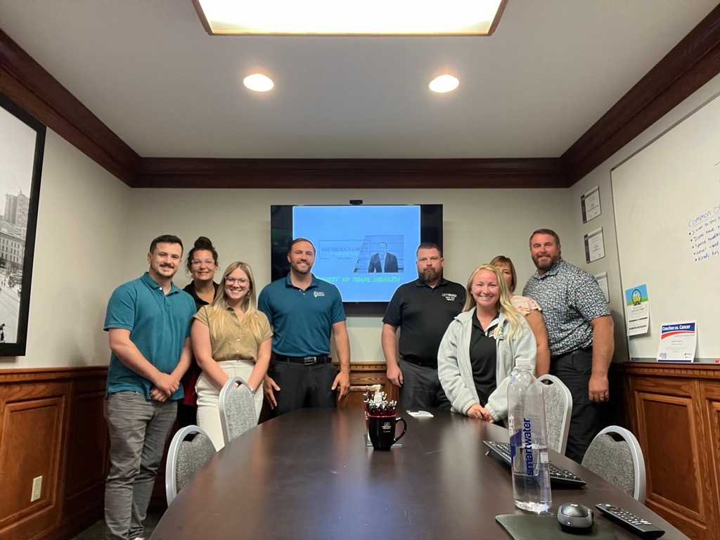 Group of eight people standing around a conference table in a room with a whiteboard and a TV screen displaying a presentation.