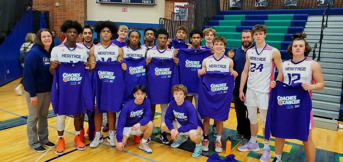 Group of young basketball players and coaches standing on a gymnasium basketball court, holding blue bags with 'Coaches vs. Cancer' logos, celebrating their participation in a game or event.