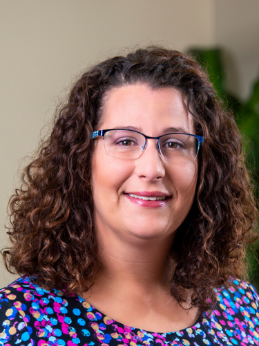 Portrait of a woman with curly brown hair, glasses, and a colorful patterned top, smiling indoors.
