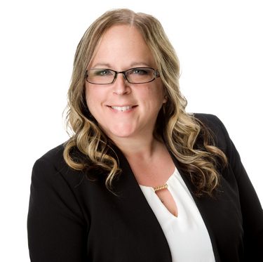 A woman with wavy blonde hair, wearing glasses, a black blazer, and a white blouse, smiling against a white background.