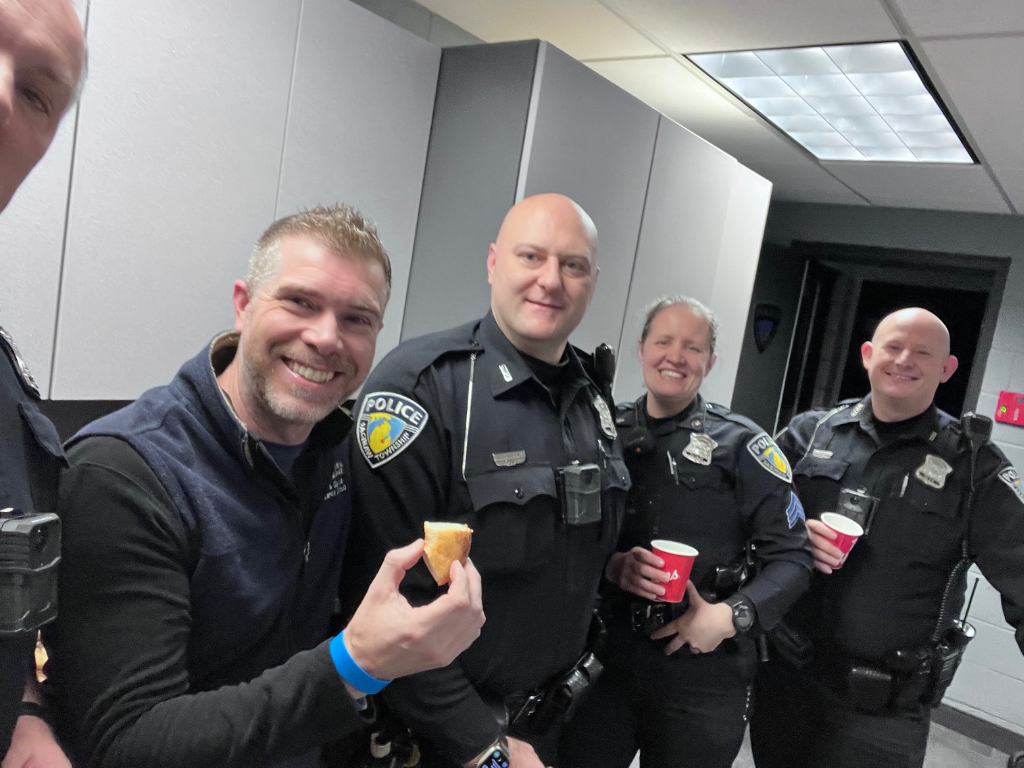 Group of five police officers smiling, two holding cups of coffee and the man in the front holding a sandwich, in a room with gray walls and ceiling lights.