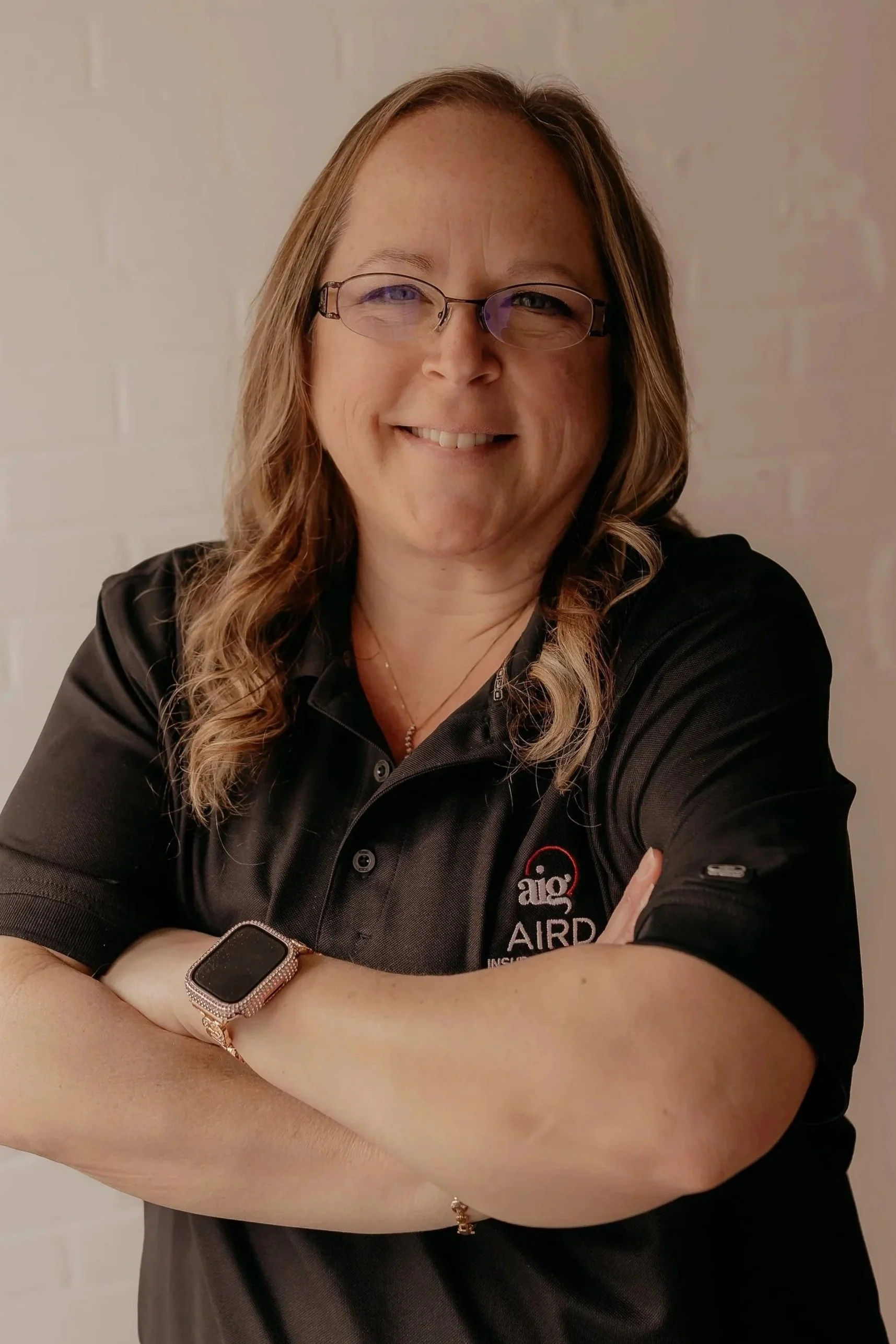 A woman with wavy blonde hair, wearing glasses, a black blazer, and a white blouse, smiling against a white background.