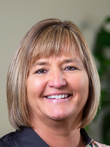 Close-up of a smiling woman with blonde hair and a black top, blurred green plant background.