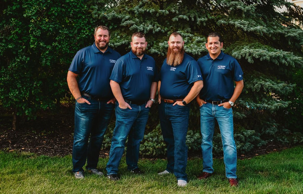 Four men standing outdoors on grass, in front of green bushes and pine trees, wearing matching blue polo shirts and jeans.