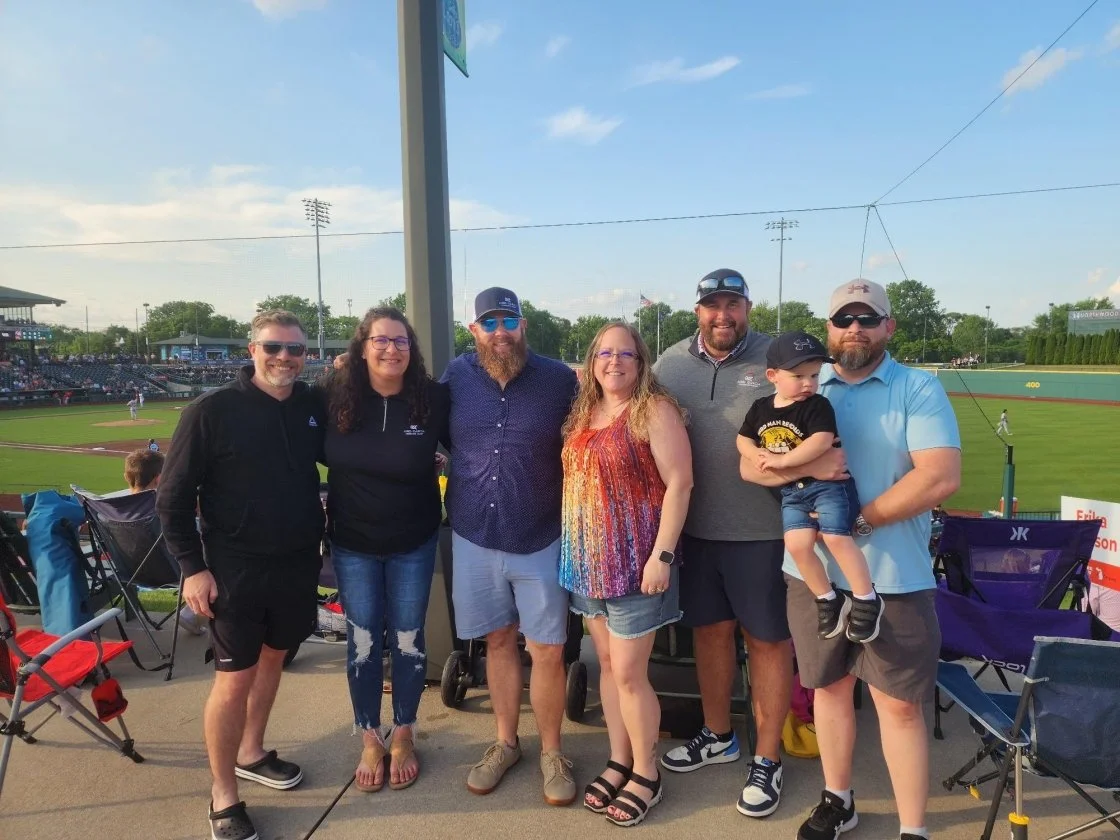 Group of six people standing in front of a baseball field, smiling at the camera. There are chairs and spectators in the background.