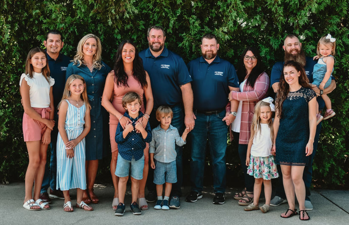 Group of adults and children standing outdoors in front of green bushes, posing for a photo.
