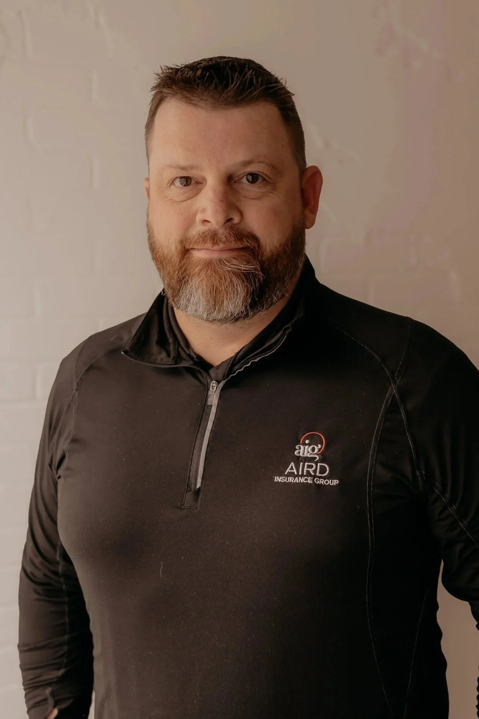 A man with short dark hair and a beard, wearing a navy blue polo shirt with the Farm Bureau Insurance logo and text that reads "Aird Garcia Insurance Group," standing in front of a light-colored brick wall.