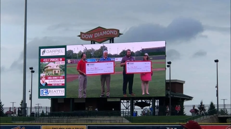 Large electronic scoreboard at a baseball stadium displaying four people holding oversized checks during an award presentation or donation event.