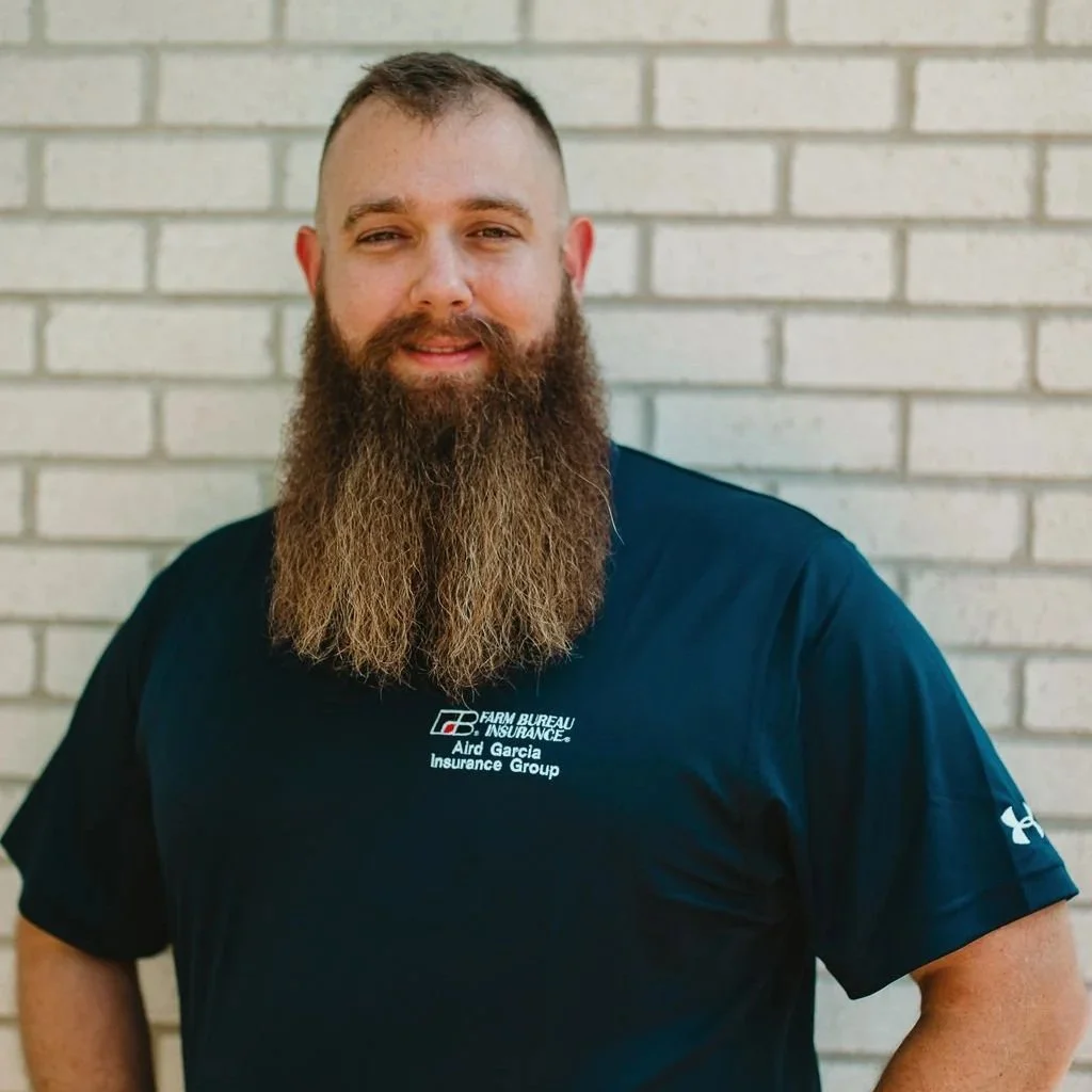 A man with a long beard and short hair standing in front of a white brick wall, wearing a dark blue sports shirt with a logo and text on it.
