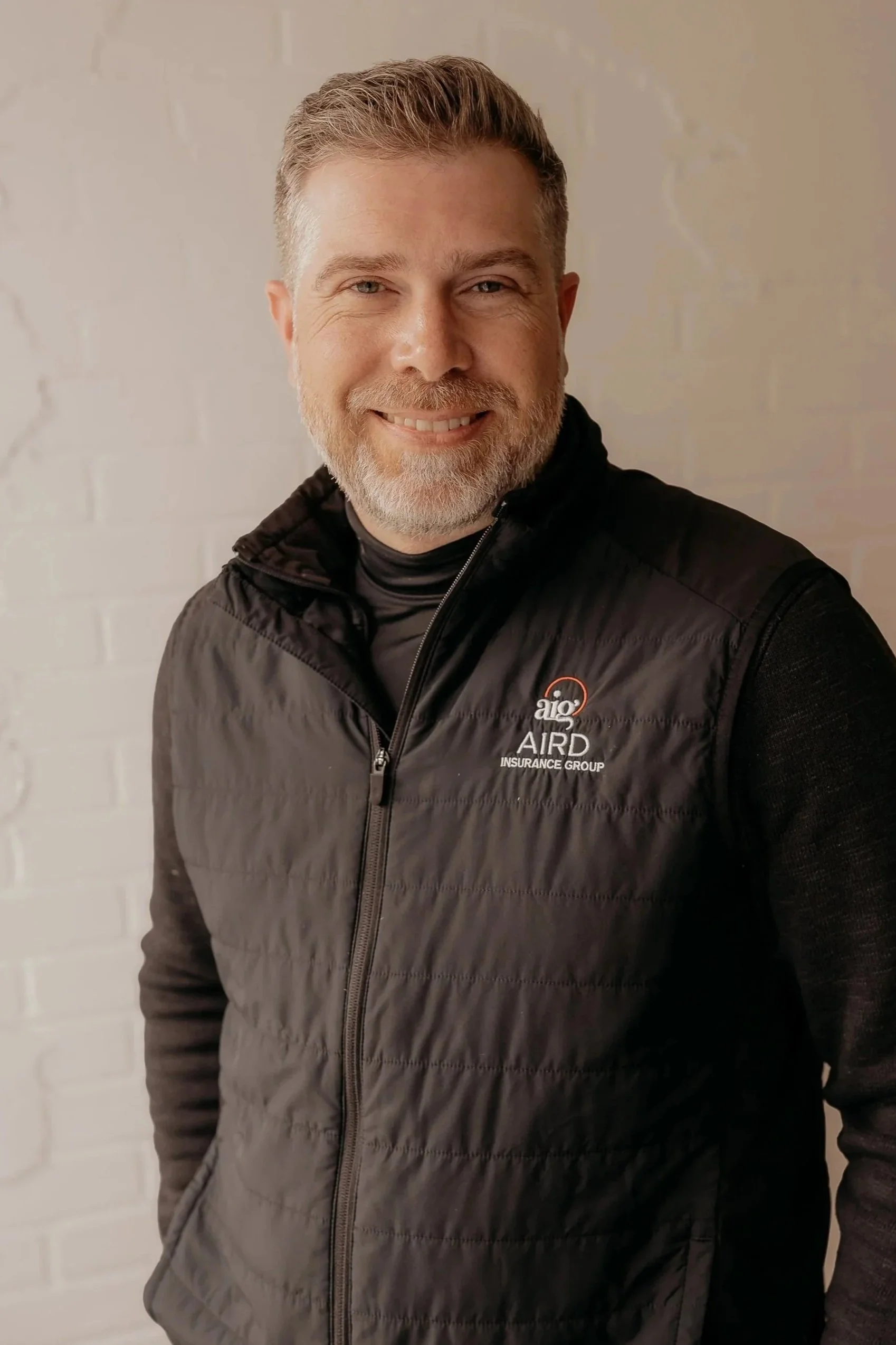 A man with short gray hair and a beard, wearing a dark blue jacket with a patch on the chest, standing against a brick wall with his arms crossed.