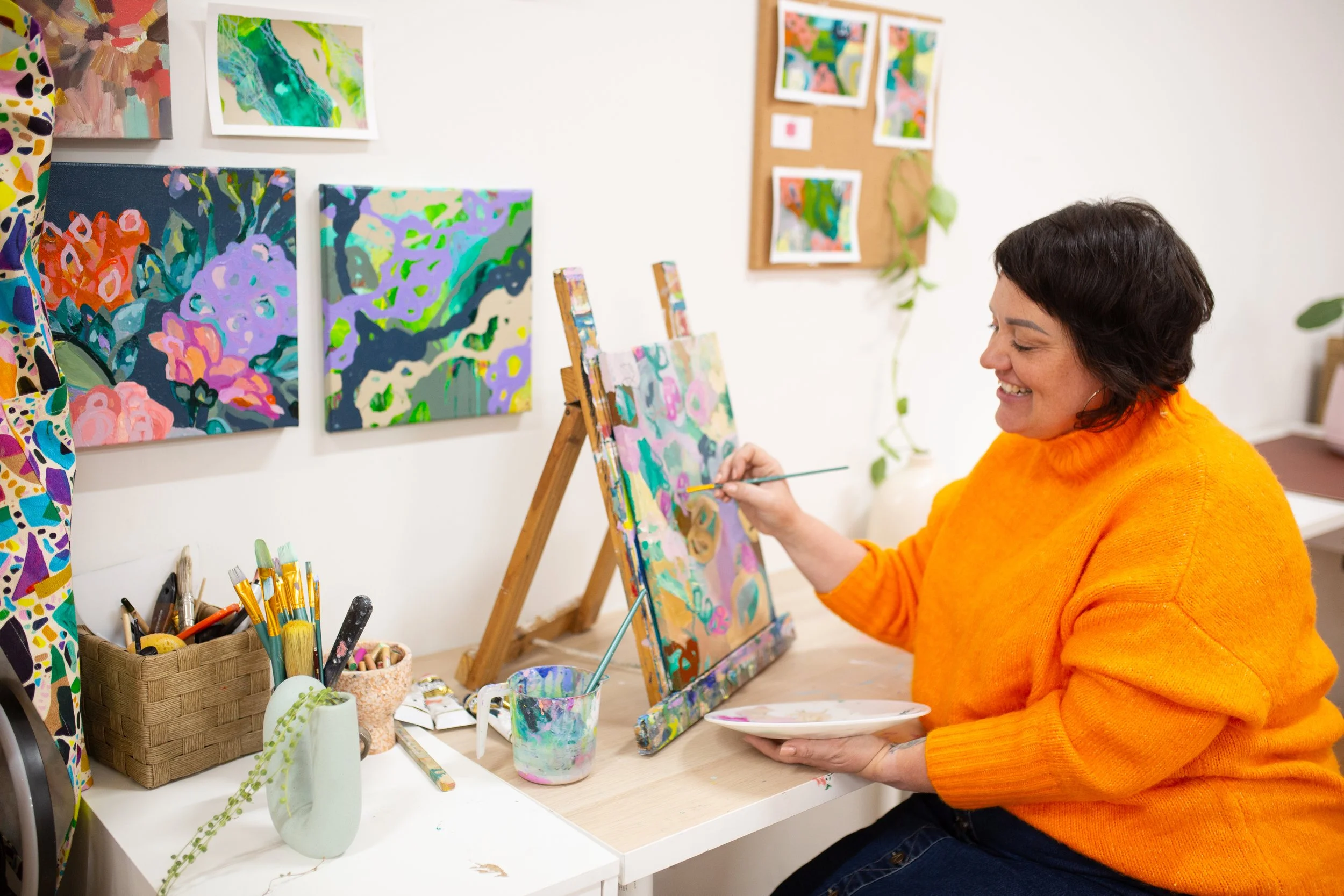 A woman in a bright orange sweater sits at a desk, painting an abstract colorful artwork on a canvas. Behind her on the wall are several other colorful paintings and a corkboard with more artwork. The desk has paintbrushes, art supplies, and a cup filled with paint. She appears happy and focused on her art.