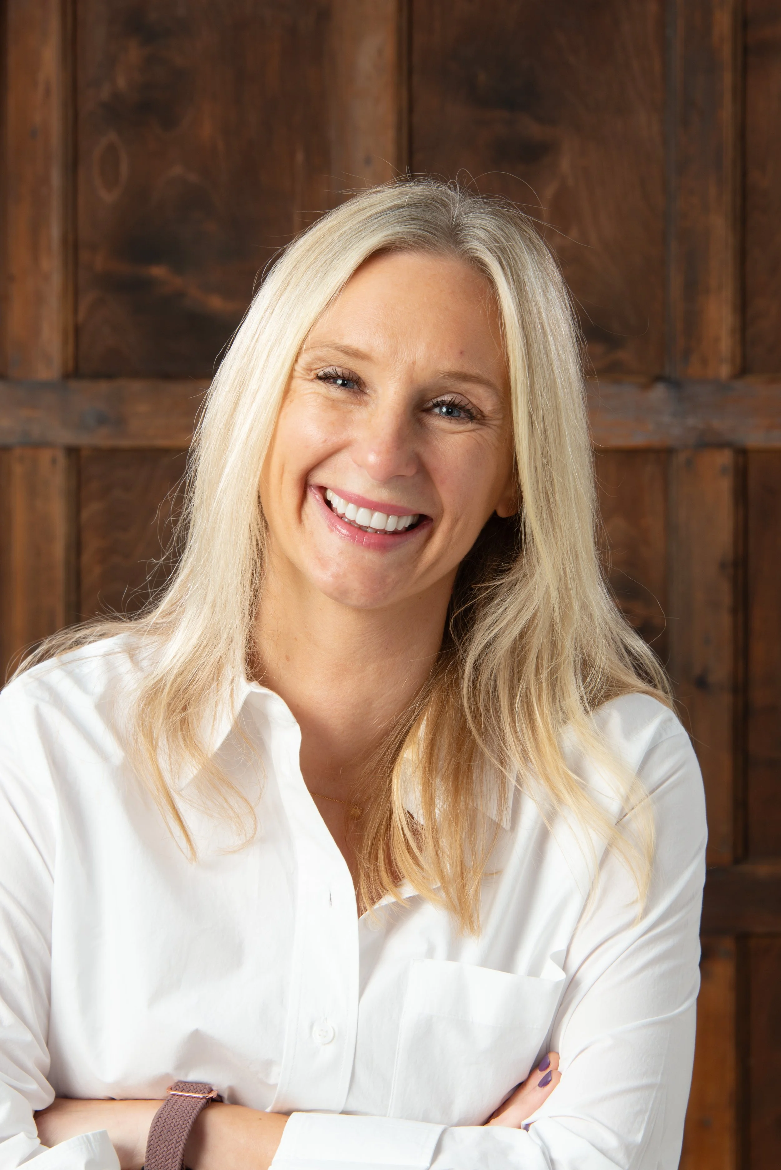 Smiling blonde woman in white shirt with crossed arms against a wooden background.