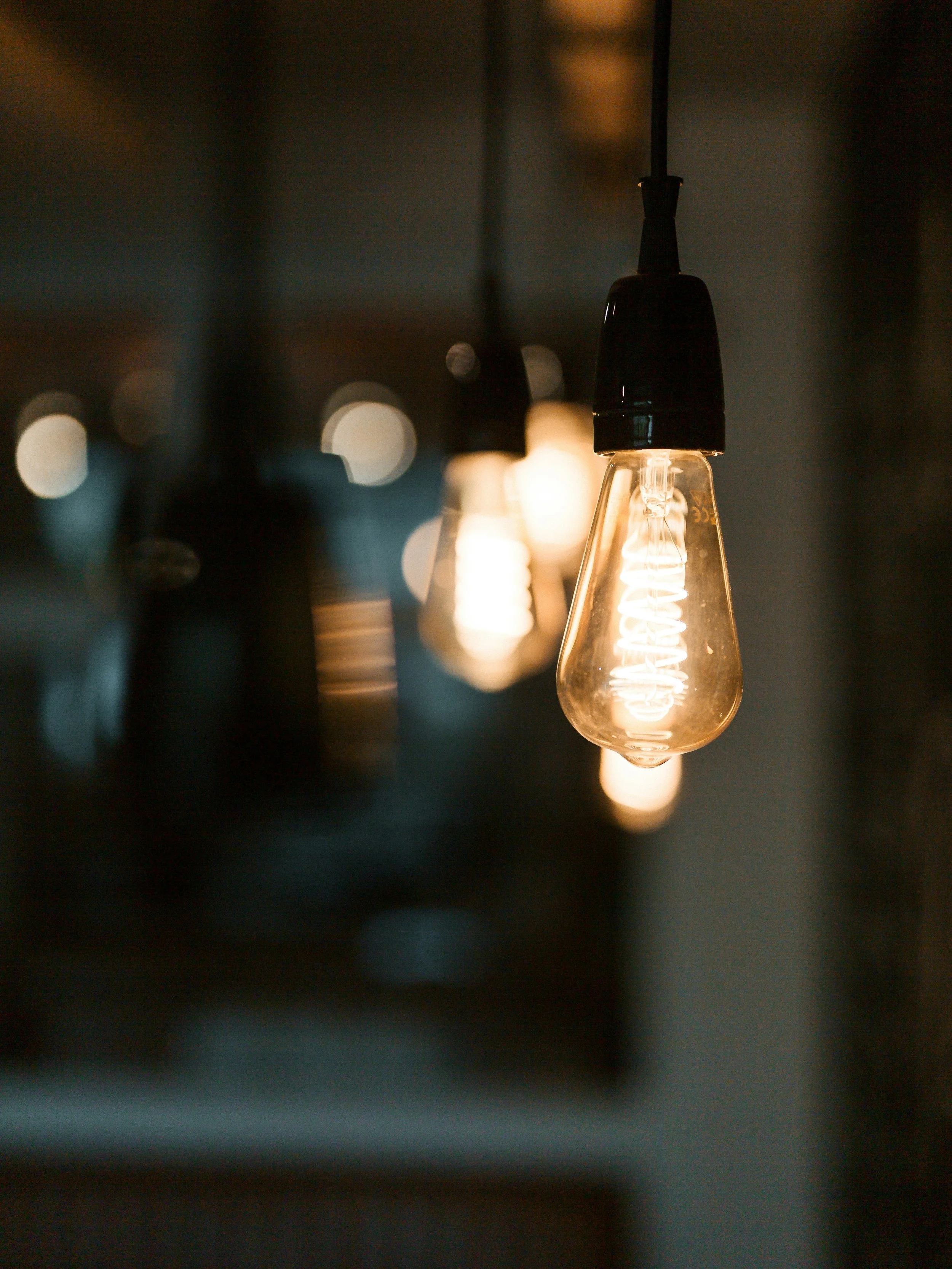Close-up of a lit Edison-style filament light bulb hanging from the ceiling with other blurred light bulbs in the background.