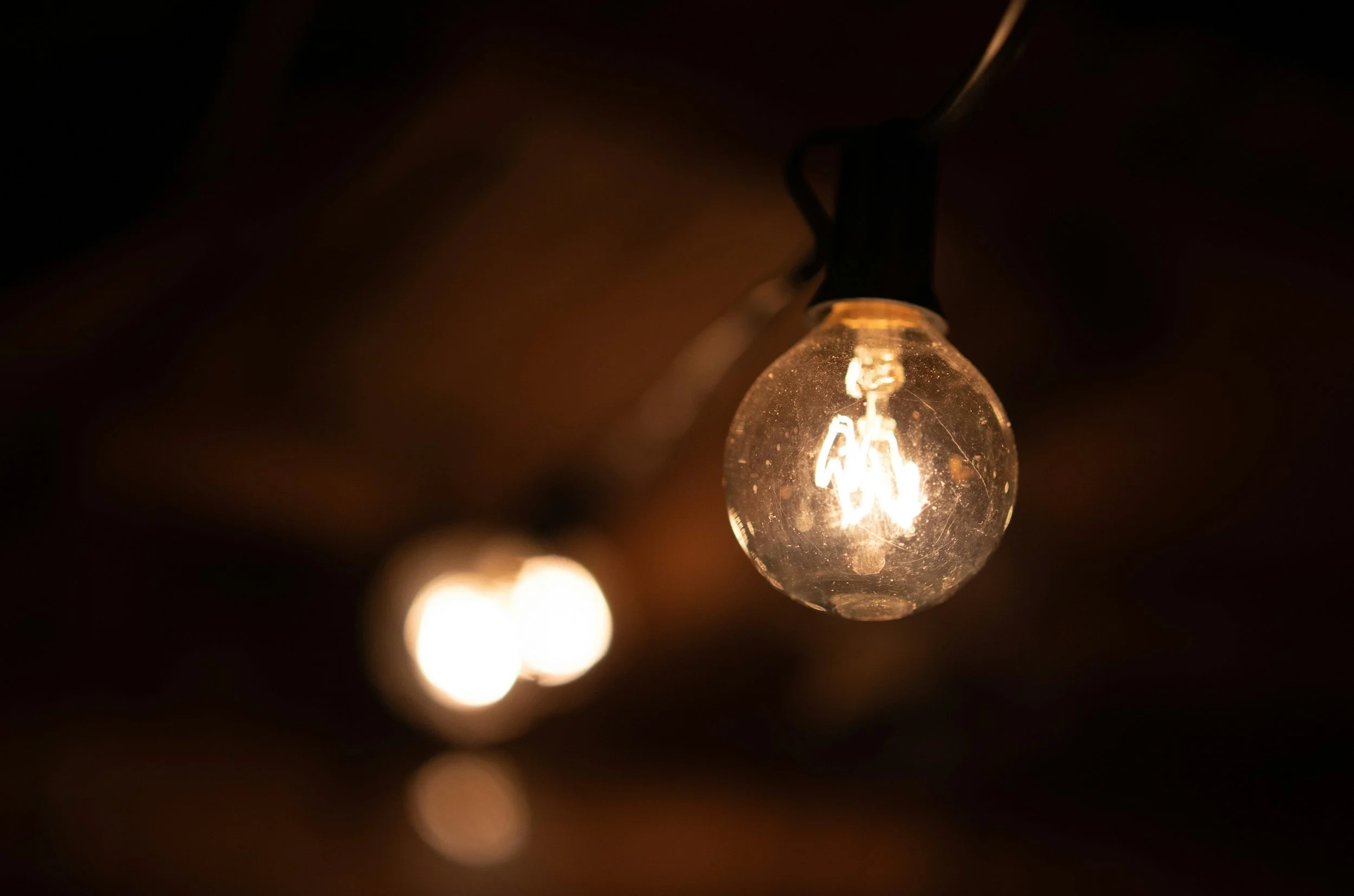 A glowing light bulb hanging from a string against a dark background, with a blurred secondary bulb nearby.