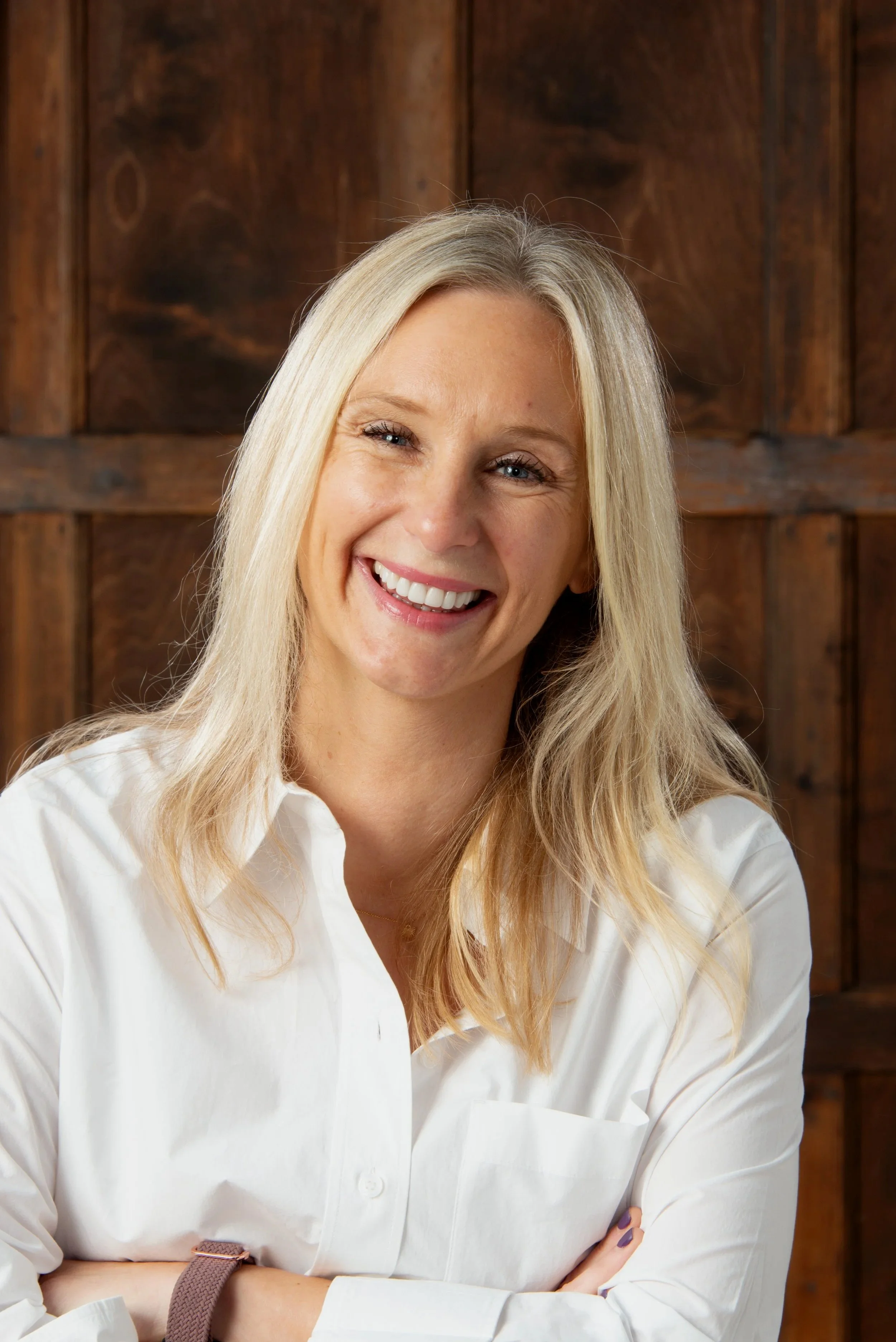 Close-up of a smiling blonde woman wearing a white shirt in front of a wooden wall.