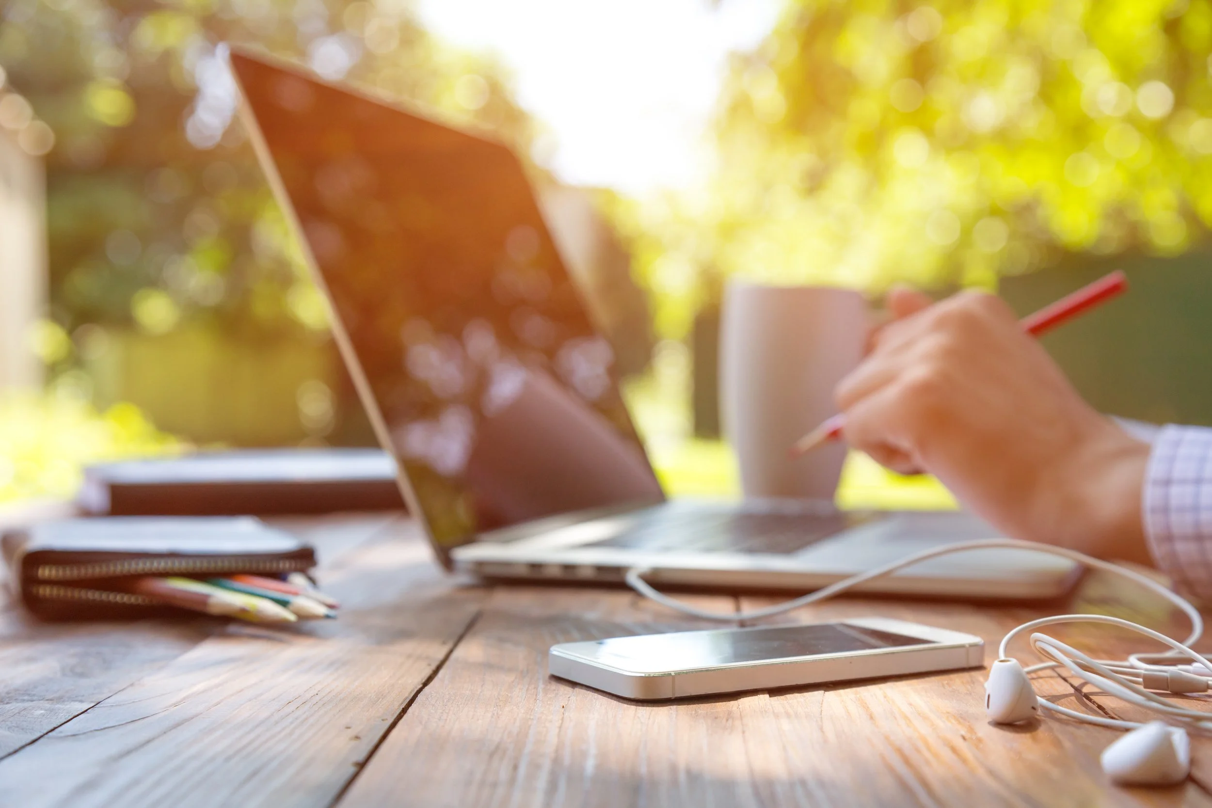 Person sitting at a wooden table using a laptop, with a smartphone, earbuds, and a mug, outdoors with sunlight and greenery in the background.
