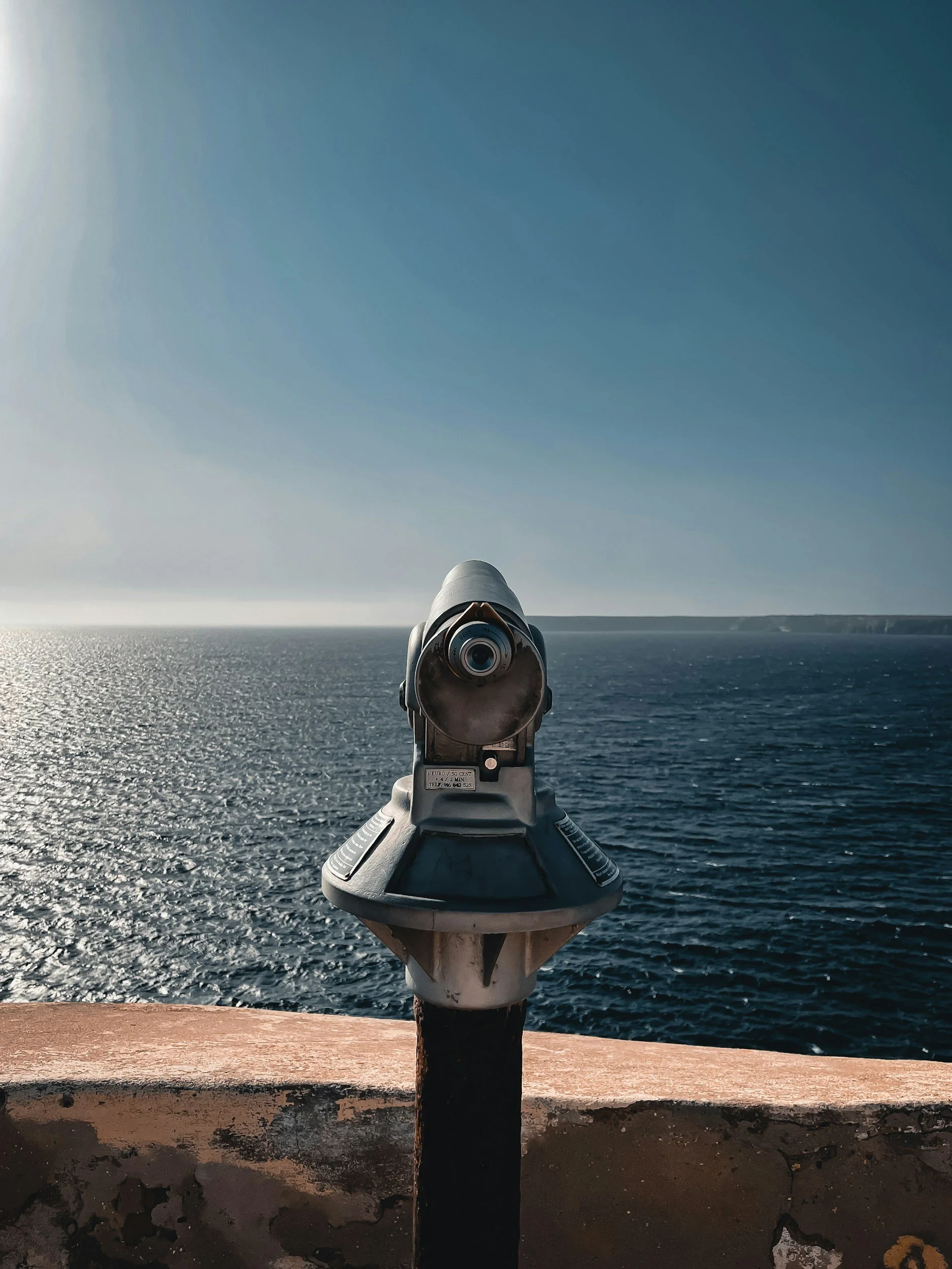 A coin-operated viewfinder on a ship deck overlooking the ocean, with the horizon and a distant landmass under a clear sky.