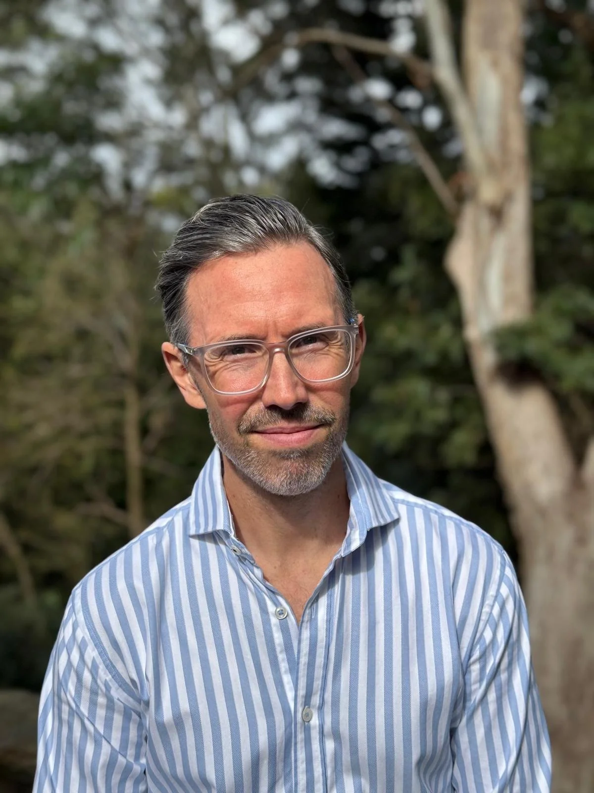 A man with gray hair, glasses, and a beard wearing a striped button-up shirt outdoors with blurred trees in the background.