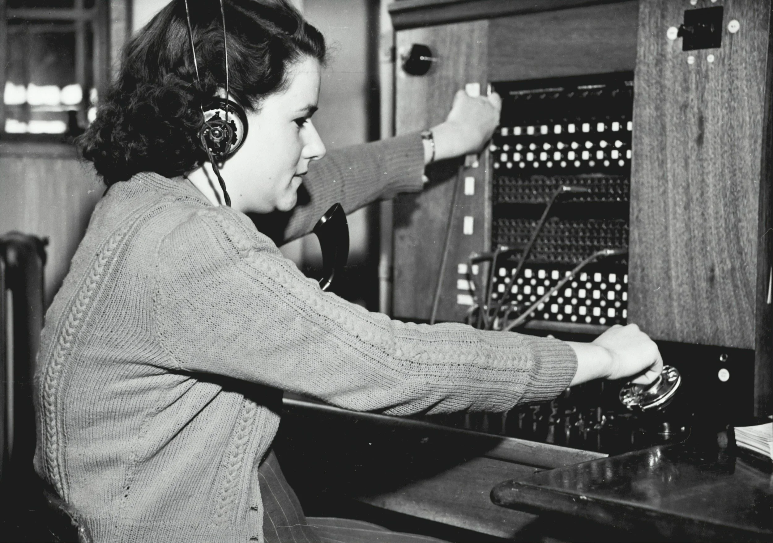 A woman with dark hair, wearing a cardigan, working at a vintage telephone switchboard with headphones.