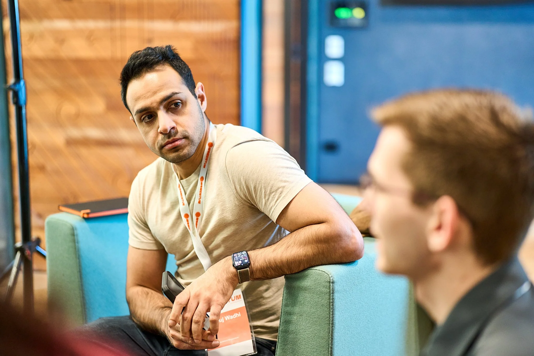A man with dark hair and a beige shirt sitting on a green sofa, looking at someone out of frame, wearing a smartwatch on his left wrist, in a room with wooden walls and a black and orange notebook on his lap.