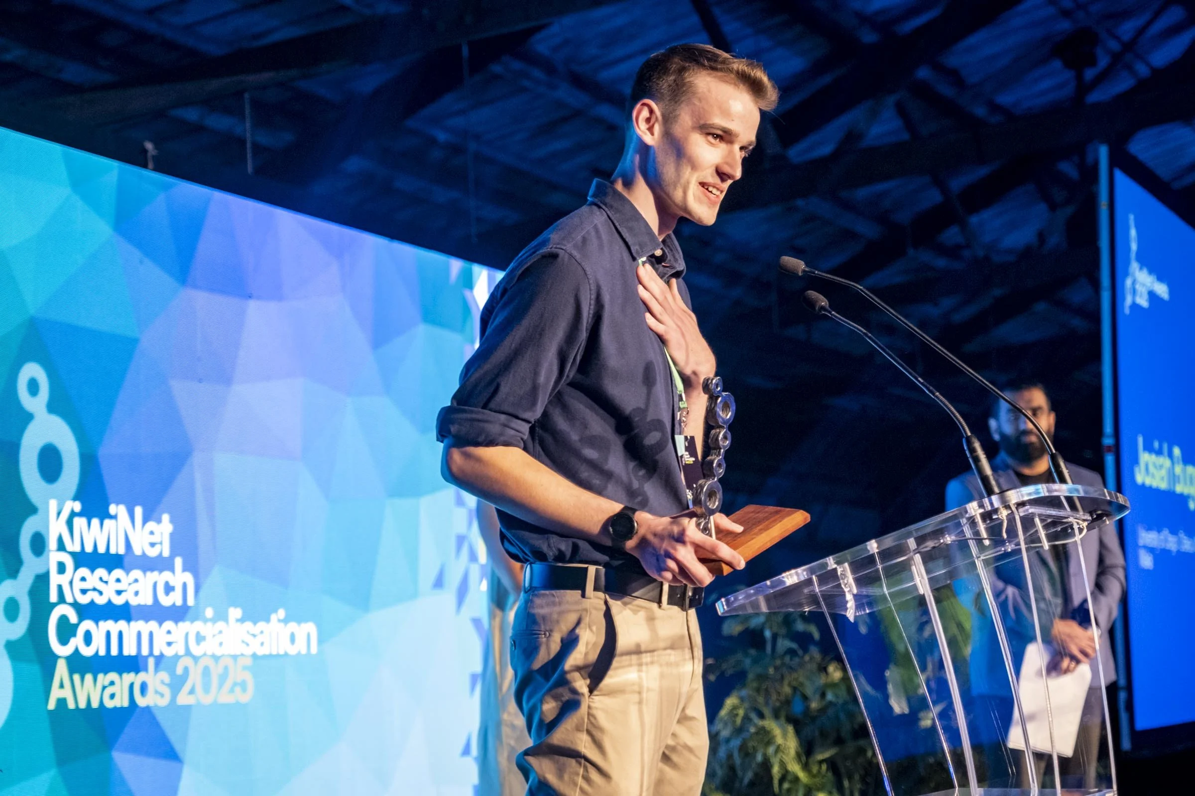 Young man with short hair holding a trophy, standing at a clear podium, smiling with his hand on his chest, during the KiwiNet Research Commercialisation Awards 2025, with a man in the background and a large blue screen displaying the event logo.