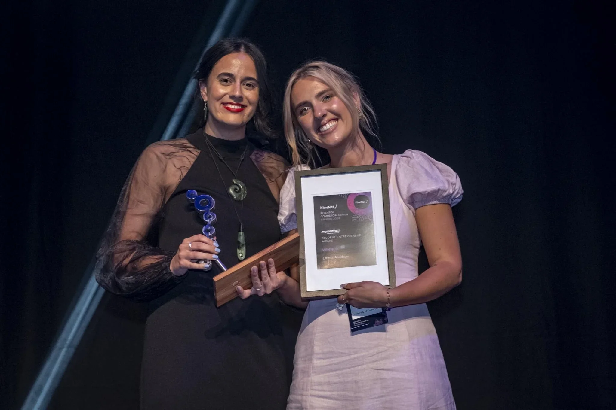 Two women on stage at an awards ceremony, one holding a trophy and the other holding a framed certificate, both smiling.