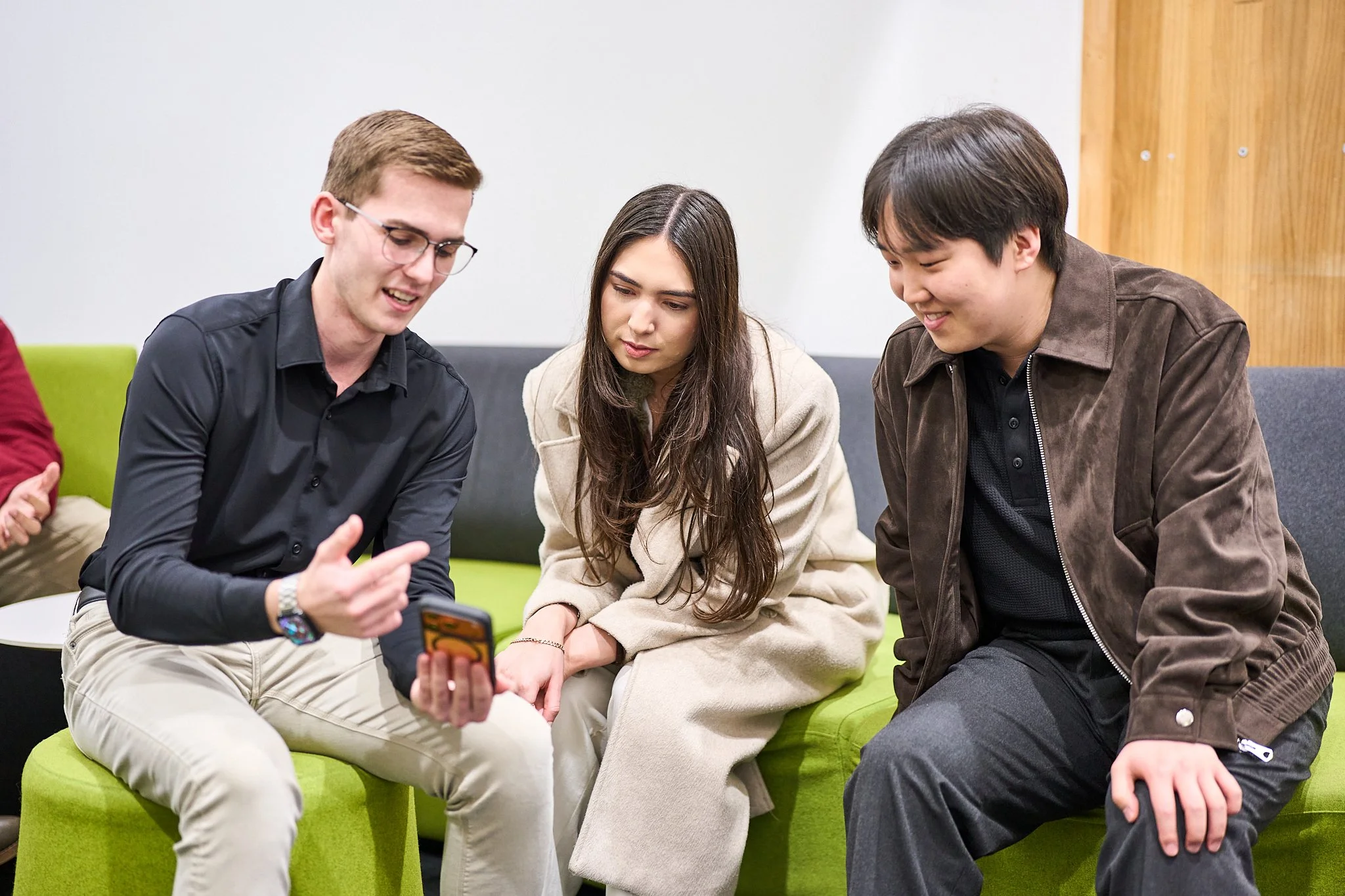Three young adults sitting on green seating, looking at a phone together, in a modern room with wooden paneling and white walls.